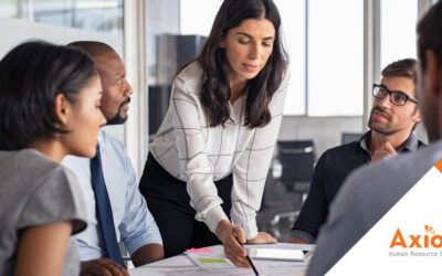 Group of diverse professionals collaborating in a meeting, with a woman leading the discussion and presenting on a document, Axiom Human Resource Solutions branding visible.