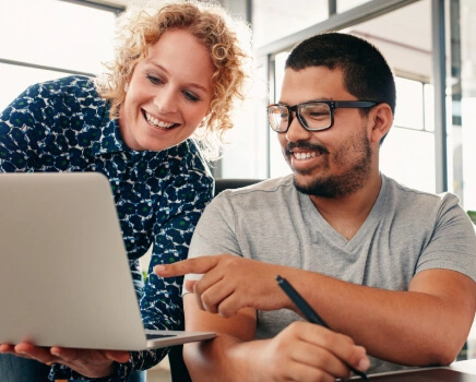 Two business partners smiling while looking at a laptop and reviewing a solution to payroll and compliance management.