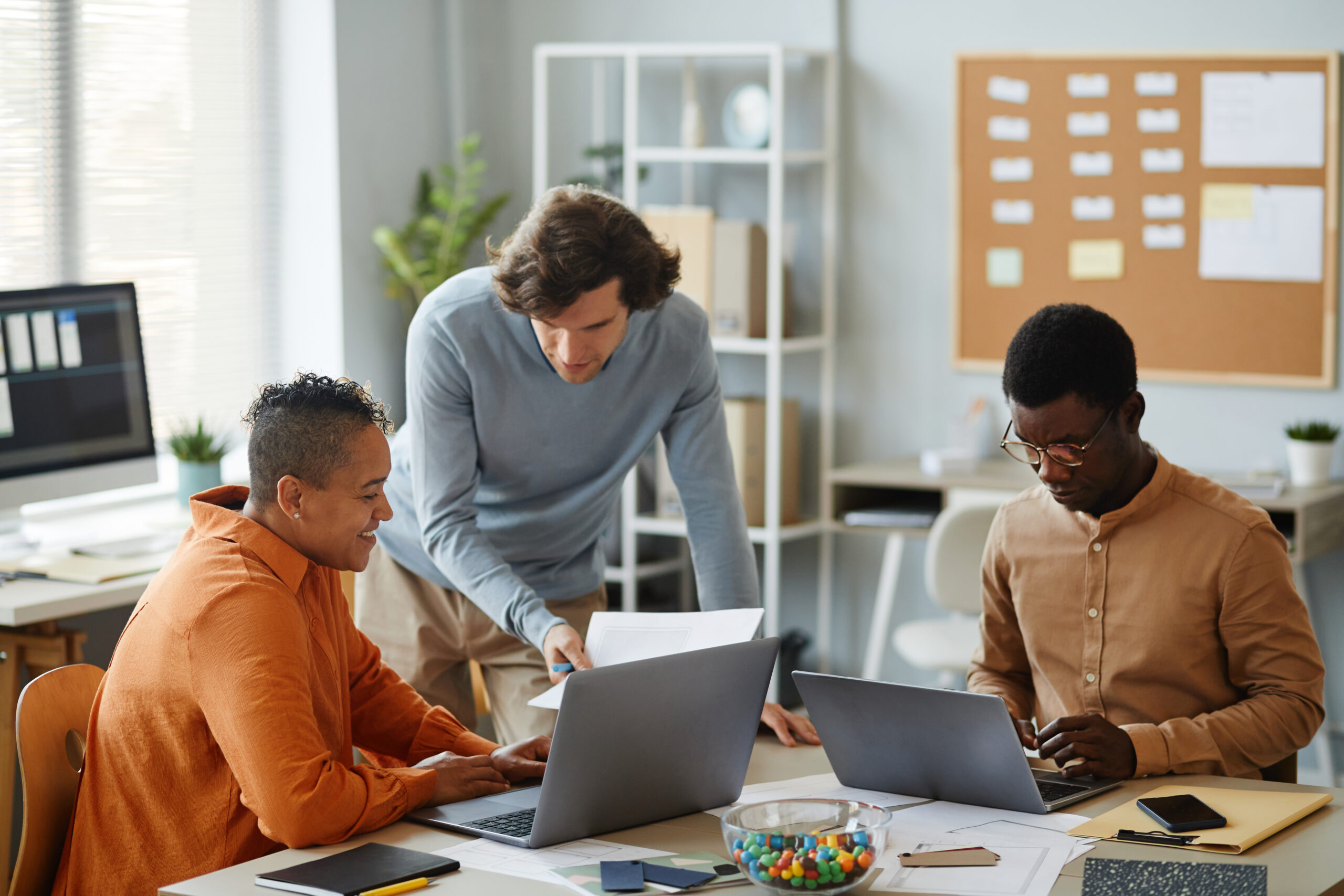 Three professionals collaborating in an office setting, reviewing documents and working on laptops, emphasizing teamwork in payroll management and error prevention.