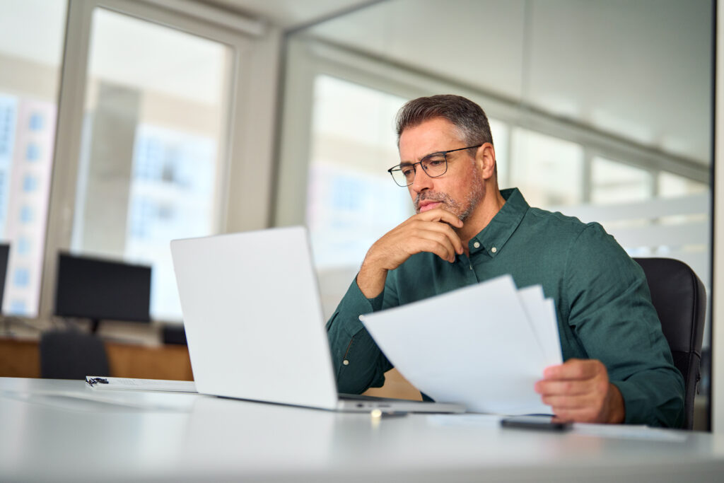 Busy middle aged HR executive, looking at laptop in office and evaluating the best payroll software options for his company.