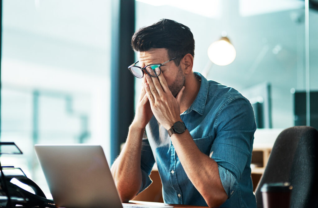 Man sitting at computer rubbing his eyes because he's frustrated over payroll mistakes.