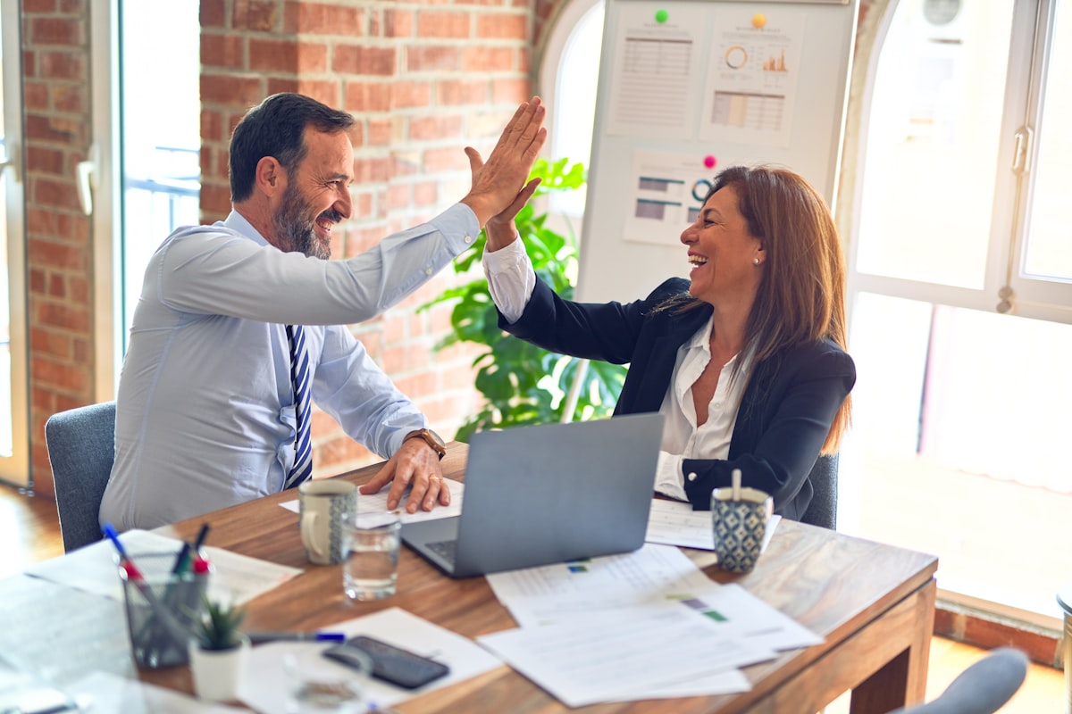 UKG Ready Partner vs Direct: 5 Critical Differences 4 UKG partner vs direct Two professionals high-fiving in a collaborative office setting, surrounded by documents and a laptop, symbolizing teamwork and successful implementation of HR solutions.